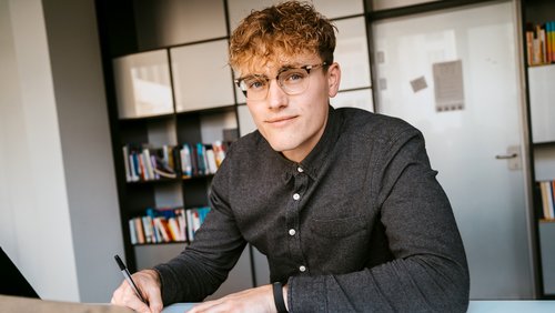 Junger Mann mit lockigem Haar und Brille, der an einem Tisch sitzt und mit einem Stift schreibt. Im Hintergrund sind Bücherregale sichtbar.
