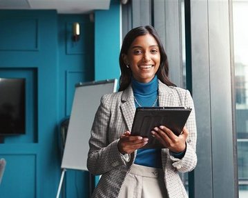 Eine Frau in einem Blazer hält ein Tablet in der Hand und lächelt, während sie in einem modernen Büro steht.