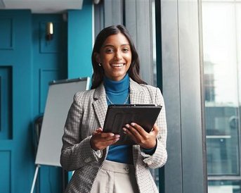A woman in a blazer is holding a tablet and smiling while standing in a modern office.