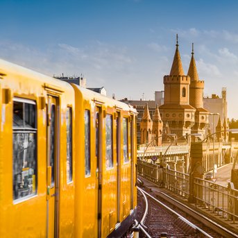 A yellow subway train travels along the tracks, with historic buildings featuring towers and a clear sky in the background.