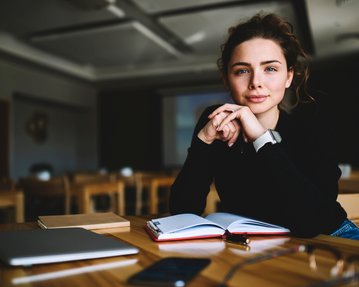 Junge Frau mit lockigem Haar sitzt an einem Tisch, schaut in die Kamera und hat ein Notizbuch sowie einen Laptop vor sich.
