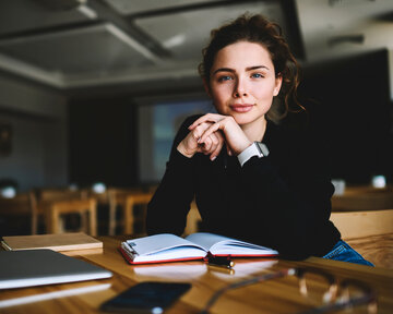 Junge Frau mit lockigem Haar sitzt an einem Tisch, schaut in die Kamera und hat ein Notizbuch sowie einen Laptop vor sich.