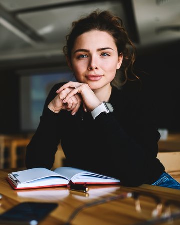 Junge Frau mit lockigem Haar sitzt an einem Tisch, schaut in die Kamera und hat ein Notizbuch sowie einen Laptop vor sich.