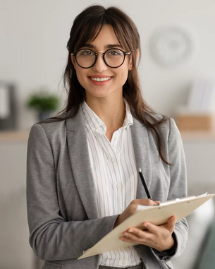 Eine Frau mit Brille trägt einen grauen Blazer und hält ein Notizbuch in der Hand. Sie steht in einem modernen Raum mit neutralen Farben und lächelt freundlich. Im Hintergrund sind Möbel und Pflanzen sichtbar.
