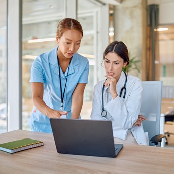 Zwei Frauen in einem modernen Büro schauen auf einen Laptop. Eine trägt einen weißen Kittel, die andere eine blaue Uniform.