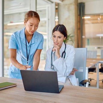 Two women in a modern office are looking at a laptop. One is wearing a white coat, while the other is in a blue uniform.