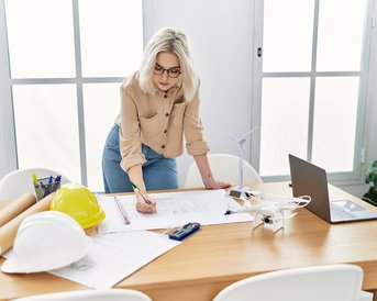 Eine blonde Frau mit Brille steht an einem Tisch und zeichnet auf Bauplänen. Neben ihr liegen Bauhelme, ein Laptop und ein Modell eines Windrads.