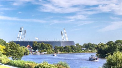 Ein modernes Stadion mit markanten Türmen, umgeben von Bäumen und einem Fluss, auf dem Boote fahren.