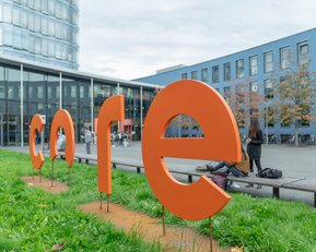 Große, orangefarbene Buchstaben bilden das Wort "core" im Freien, umgeben von Gras und einem modernen Gebäude im Hintergrund.