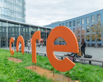 Large, orange letters spell out the word "core" outdoors, surrounded by grass and a modern building in the background.