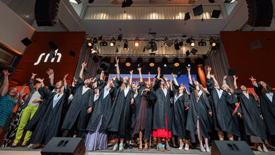 Graduates in academic gowns throw their hats during a graduation ceremony on a stage.
