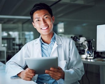 Ein junger Mann in einem Laborkittel hält ein Tablet in der Hand und lächelt. Im Hintergrund sind wissenschaftliche Geräte und Computer sichtbar.