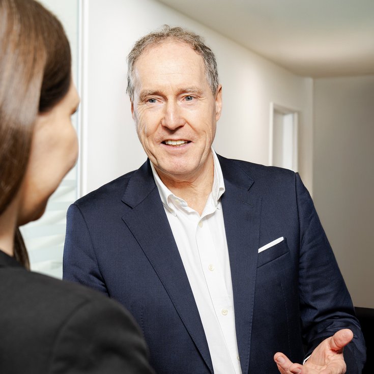 Ein Mann in einem Anzug spricht mit einer Person, die ihm zugewandt ist. Der Mann hat graue Haare und lächelt freundlich.