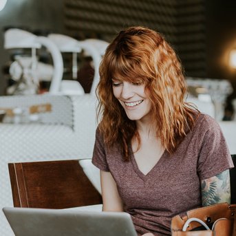Eine lächelnde Frau mit lockigem, rotem Haar sitzt an einem Tisch in einem Café und arbeitet an einem Laptop. Im Hintergrund sind verschwommene Café-Möbel und eine Neonbeleuchtung sichtbar.