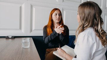 Zwei Frauen führen ein Gespräch an einem Tisch. Eine Frau mit roten Haaren spricht, während die andere Notizen macht. Ein Glas Wasser steht auf dem Tisch.