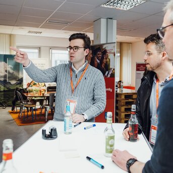 Drei Männer stehen an einem Tisch, einer zeigt mit dem Finger auf etwas. Alle tragen Namensschilder und diskutieren. Getränke sind auf dem Tisch.