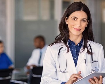 Ärztin mit Stethoskop hält ein Tablet in der Hand, lächelt und steht in einem modernen medizinischen Umfeld. Im Hintergrund sind Patienten zu sehen.