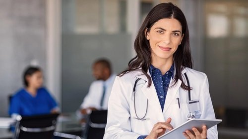 Ärztin mit Stethoskop hält ein Tablet in der Hand, lächelt und steht in einem modernen medizinischen Umfeld. Im Hintergrund sind Patienten zu sehen.
