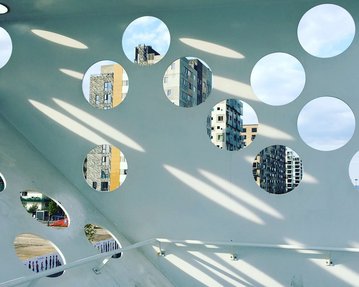 Round window openings in a modern wall offer views of urban buildings and the sky. Rays of light stream through the openings.