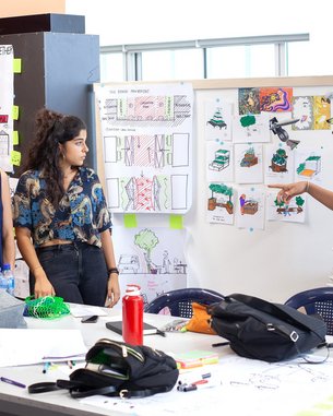 Sechs Personen stehen um einen Tisch, während eine Person eine Präsentation hält. An der Wand sind verschiedene Zeichnungen und Pläne angebracht. Einige Teilnehmer machen Notizen oder schauen aufmerksam zu.