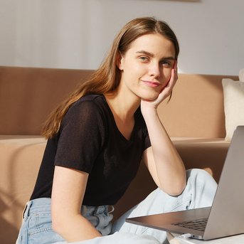 A young woman sits smiling on the floor in front of a laptop, with a cozy sofa in the background. She is wearing a black t-shirt and jeans, looking relaxed.