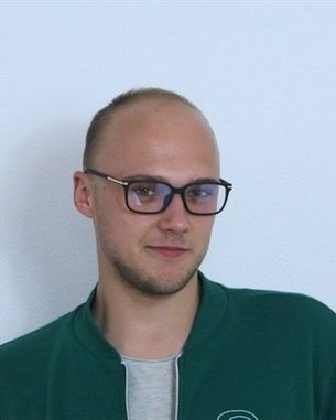 Ein Mann mit Glatze und Brille trägt eine grüne Jacke und schaut direkt in die Kamera vor einer hellen Wand.