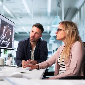 Two people are sitting at a table, looking at graphs on several monitors in a modern office.