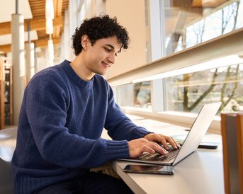 Ein junger Mann mit lockigem Haar sitzt an einem Tisch in einer Bibliothek und arbeitet konzentriert an einem Laptop.
