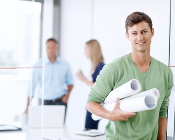 Ein junger Mann hält mehrere Pläne in der Hand und lächelt, während zwei Personen im Hintergrund miteinander sprechen.
