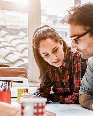 Drei Personen arbeiten gemeinsam an einem Tisch, während sie Notizen machen und Ideen austauschen. Eine Person trägt eine Mütze, eine andere hat eine karierten Hemd an, und die dritte trägt eine Brille.