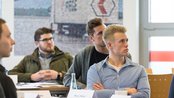 Eine Gruppe von jungen Männern sitzt an einem Tisch und hört aufmerksam zu, während eine Präsentation im Hintergrund stattfindet.