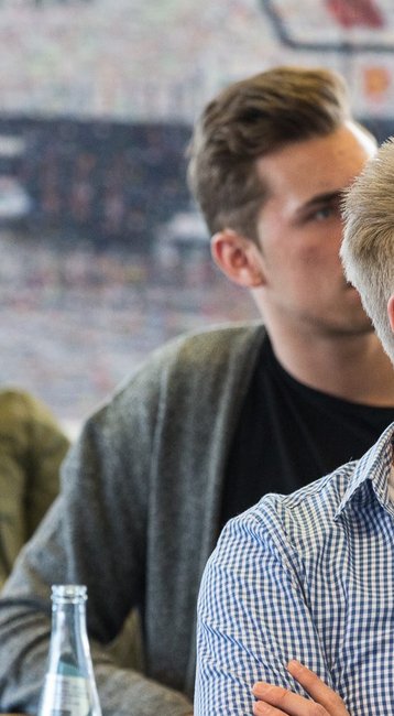 Eine Gruppe von jungen Männern sitzt an einem Tisch und hört aufmerksam zu, während eine Präsentation im Hintergrund stattfindet.