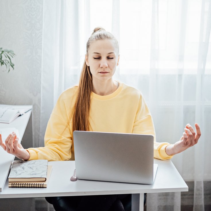 A woman in a yellow sweatshirt sits at a desk, meditating while sitting in front of a laptop. Plants are in the background.