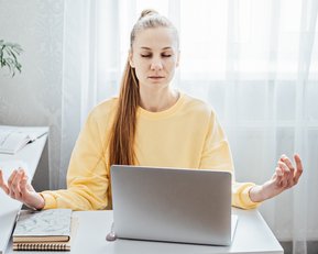 A woman in a yellow sweatshirt sits at a desk, meditating while sitting in front of a laptop. Plants are in the background.