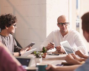 Eine Gruppe von Personen sitzt an einem Tisch in einem Besprechungsraum. Ein Mann mit Brille spricht, während andere Teilnehmer Notizen machen oder Tablets verwenden. Pflanzen und Getränke sind auf dem Tisch verteilt.