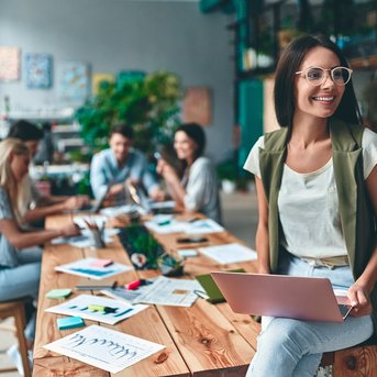 A woman with glasses is sitting on a table working on a laptop, while several people in the background are working on a creative project.