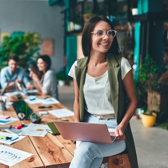 A woman with glasses is sitting on a table working on a laptop, while several people in the background are working on a creative project.