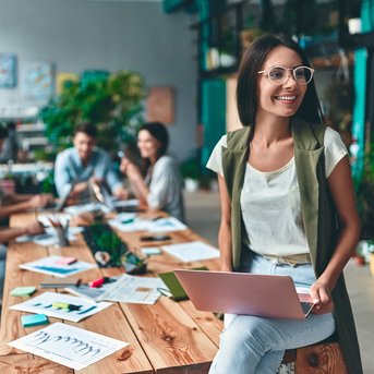 Eine Frau mit Brille sitzt auf einem Tisch und arbeitet an einem Laptop, während mehrere Personen im Hintergrund an einem kreativen Projekt arbeiten.