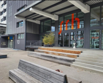 Entrance of a modern building with large windows and a striking red sign reading 'srh'. In front of the building, there are wooden steps and a bench.