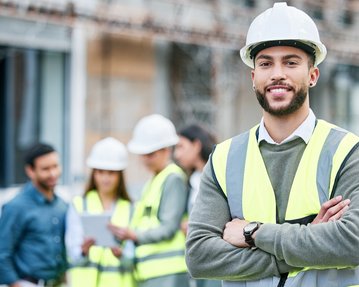 A construction manager in safety gear and a helmet stands smiling in the foreground, while in the background, other workers are engaged in a project.