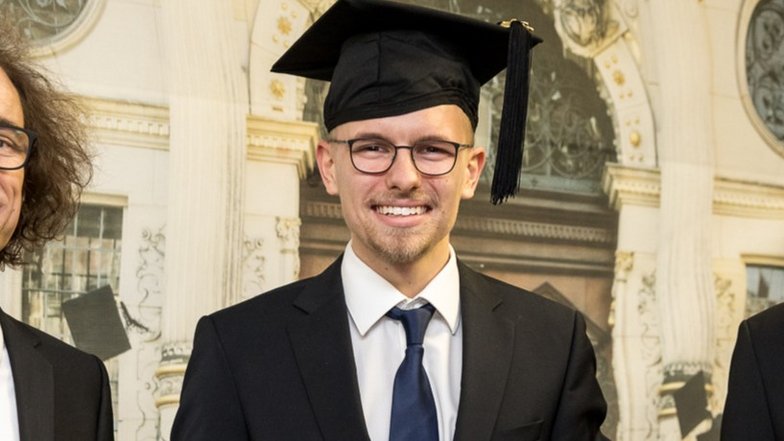 Ein graduierter Mann trägt eine akademische Mütze und einen Anzug, lächelt und steht neben zwei anderen Personen.
