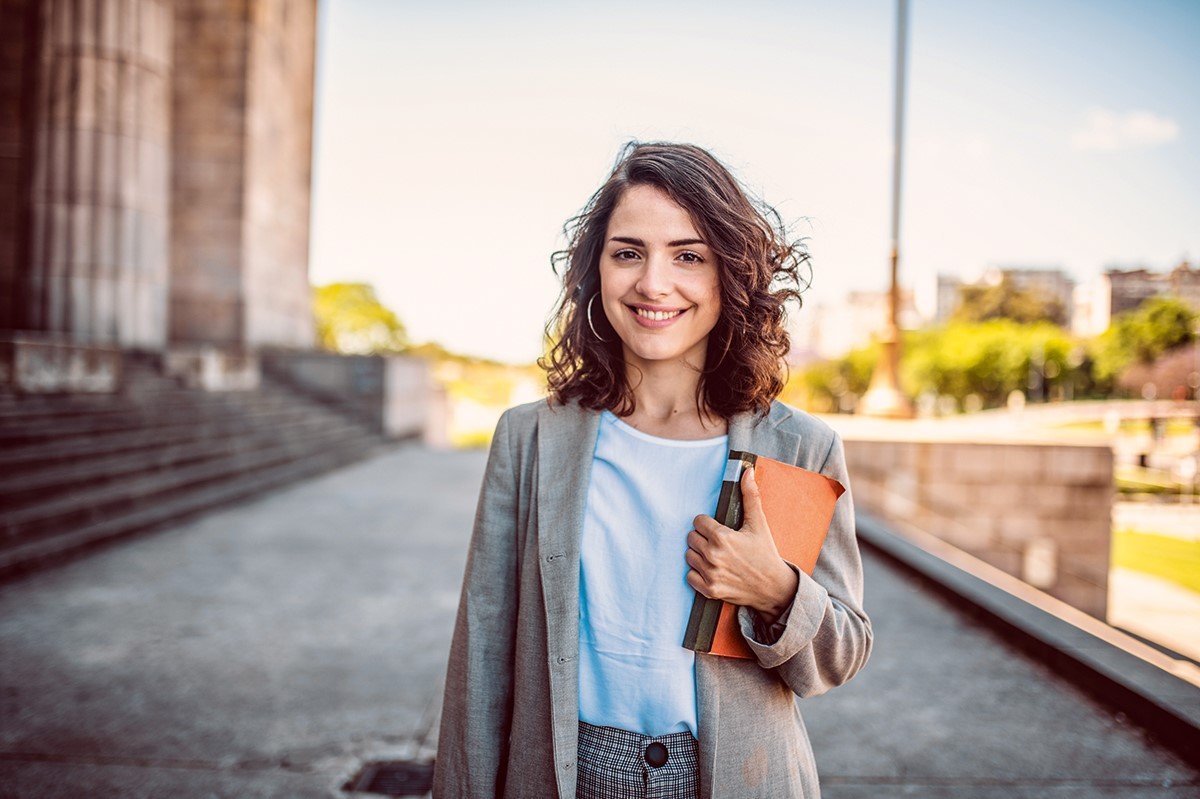 Eine junge Frau mit lockigem Haar trägt einen Blazer und hält ein Notizbuch sowie einen Ordner. Sie steht vor einem architektonischen Hintergrund und lächelt freundlich in die Kamera.