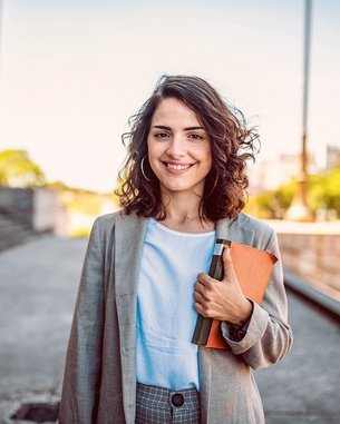 Eine junge Frau mit lockigem Haar trägt einen Blazer und hält ein Notizbuch sowie einen Ordner. Sie steht vor einem architektonischen Hintergrund und lächelt freundlich in die Kamera.