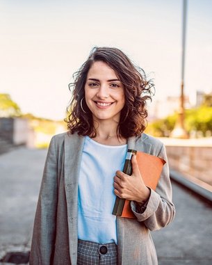 Eine junge Frau mit lockigem Haar trägt einen Blazer und hält ein Notizbuch sowie einen Ordner. Sie steht vor einem architektonischen Hintergrund und lächelt freundlich in die Kamera.