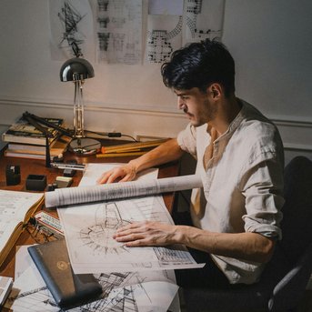 Ein junger Mann sitzt an einem Tisch, studiert Baupläne und Zeichnungen, umgeben von Werkzeugen und Büchern, während eine Lampe Licht auf seine Arbeit wirft.