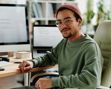 Ein junger Mann mit Brille und rotem Hut sitzt an einem Schreibtisch. Er trägt ein grünes Sweatshirt und lächelt. Im Hintergrund sind Computerbildschirme und Büromaterial sichtbar.