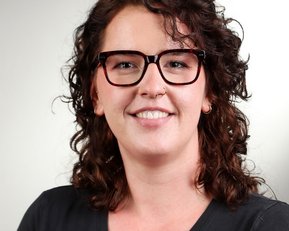 Eine lächelnde Frau mit lockigem, braunem Haar und Brille, die ein schwarzes Oberteil trägt, posiert vor einem hellen Hintergrund.
