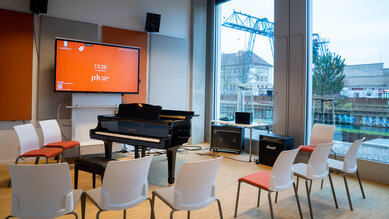 A room with a piano, chairs, and a screen. Large windows offer a view of a bridge and water. The room is bright and furnished in a modern style.