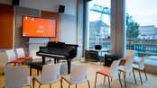 A room with a piano, chairs, and a screen. Large windows offer a view of a bridge and water. The room is bright and furnished in a modern style.