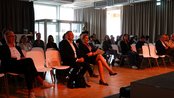 Eine Gruppe von Menschen sitzt in einem Konferenzraum auf weißen Stühlen. Einige Teilnehmer hören aufmerksam zu, während andere auf die Bühne blicken. Der Raum ist gut beleuchtet und hat große Fenster.